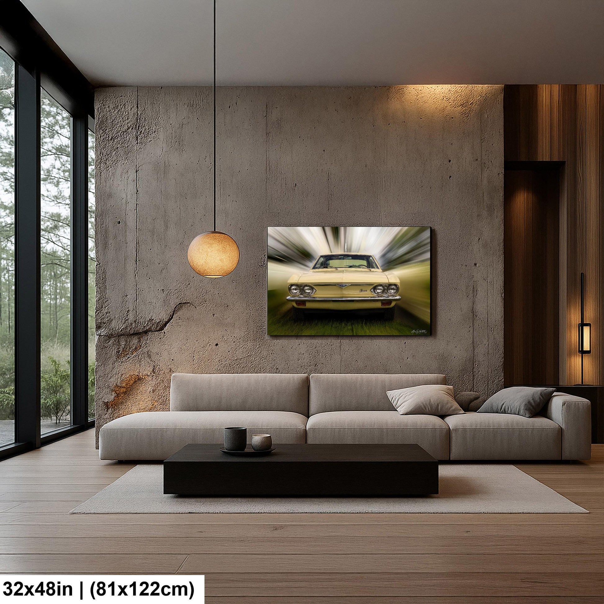 a modern living room with a large window overlooking a forested area. The room features a gray sofa, a black coffee table, and a framed artwork of a yellow car on the wall.