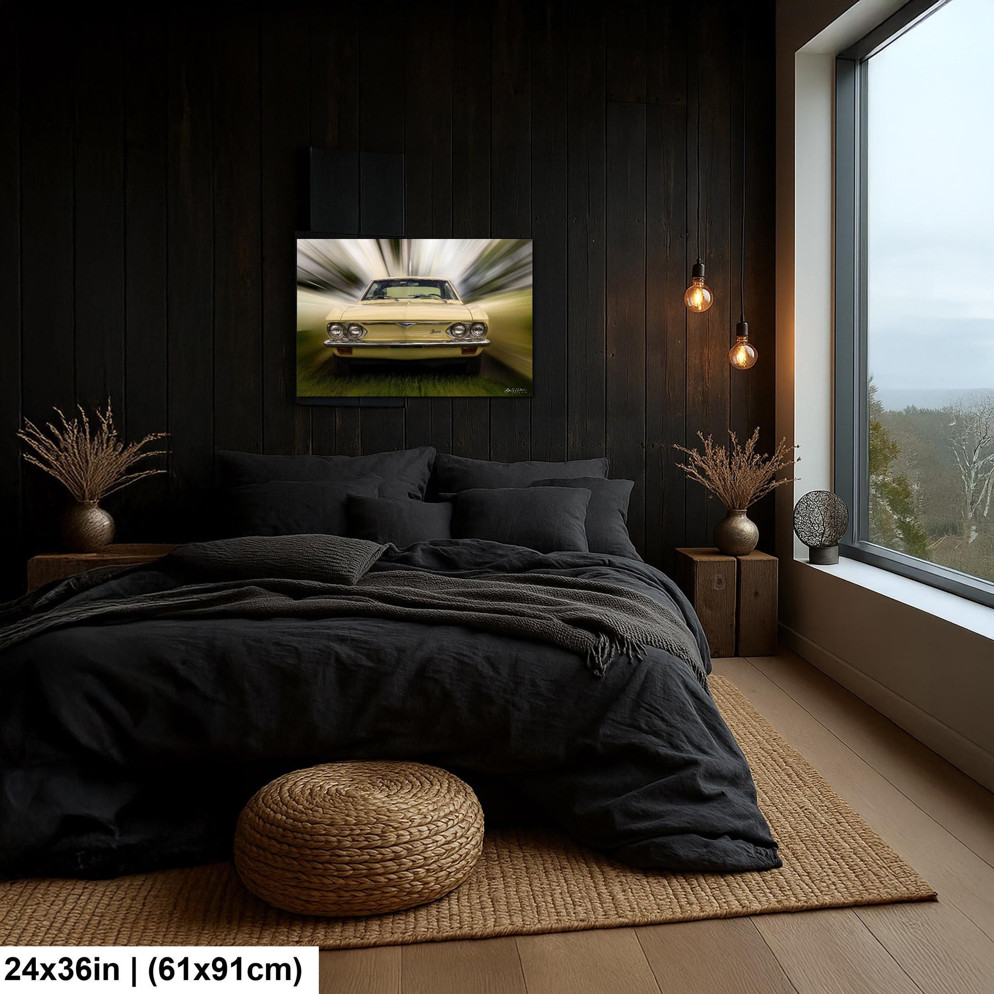 a bedroom with a large window overlooking a scenic outdoor view. The room features a black bed with a gray blanket, a woven ottoman, and a framed picture of a yellow car on the wall.
